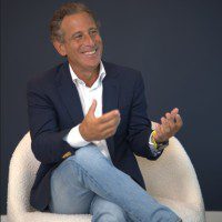 Man in blazer and jeans sitting and gesturing while smiling.