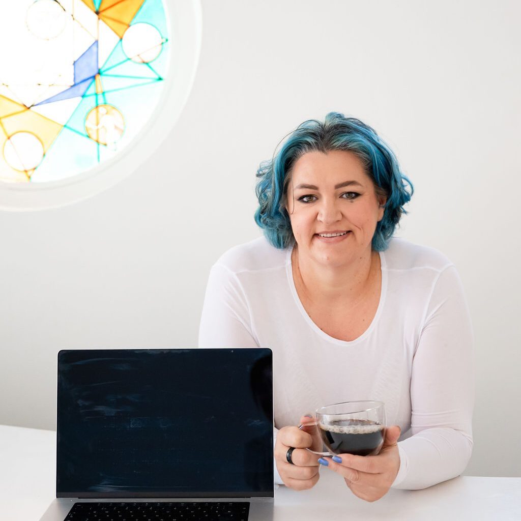 Smiling woman with blue hair holding a cup beside a laptop.